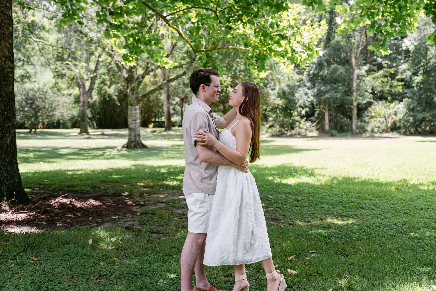 Couple sharing a romantic kiss in a lush green park, man in beige outfit and woman in white dress.