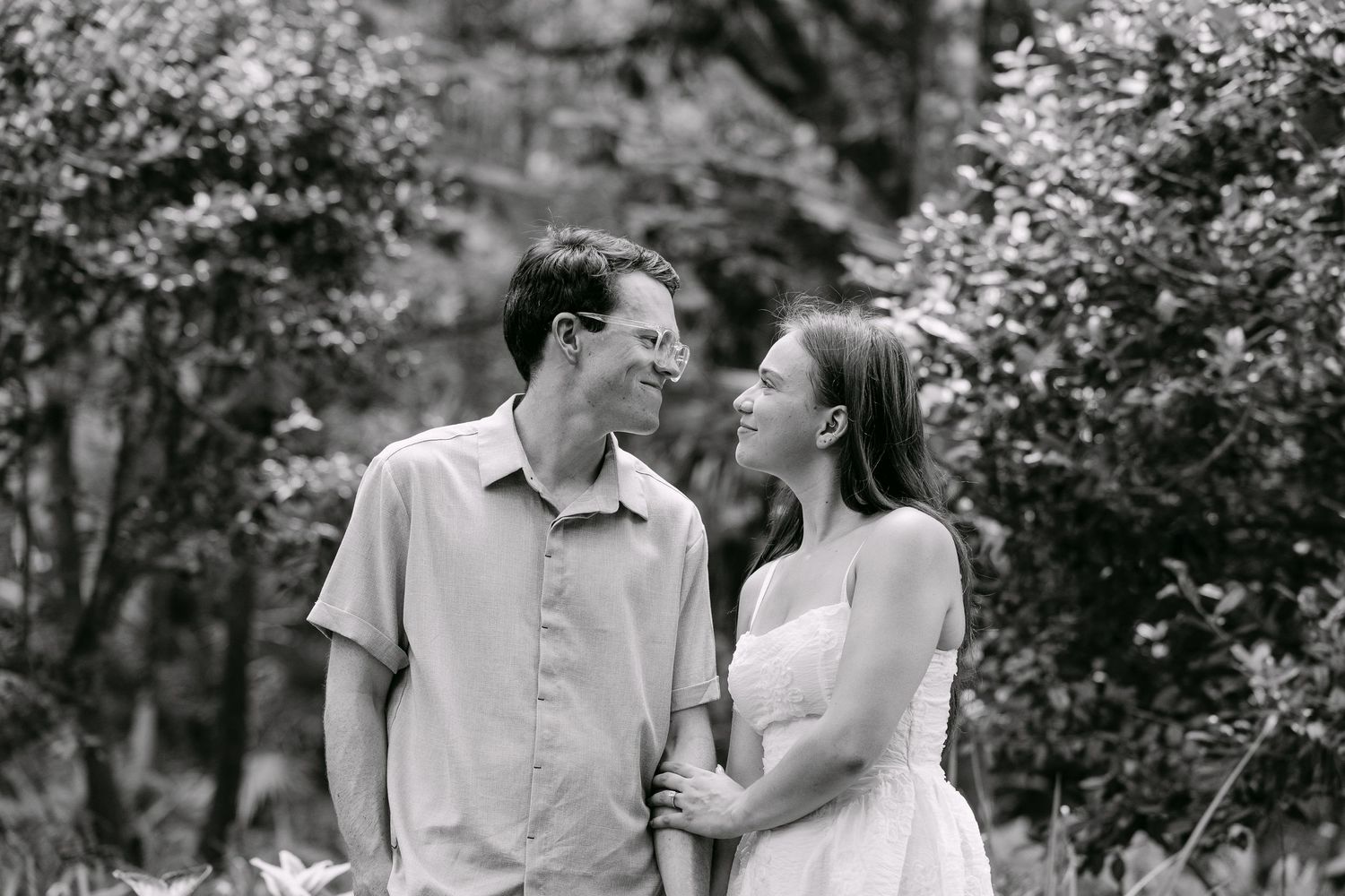 Black and white photo of couple smiling at each other surrounded by trees.