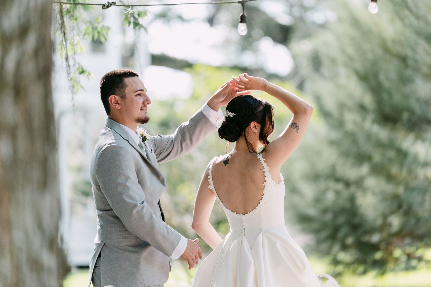 Bride and groom share first dance outdoors under string lights, groom spinning bride in white backless wedding dress.