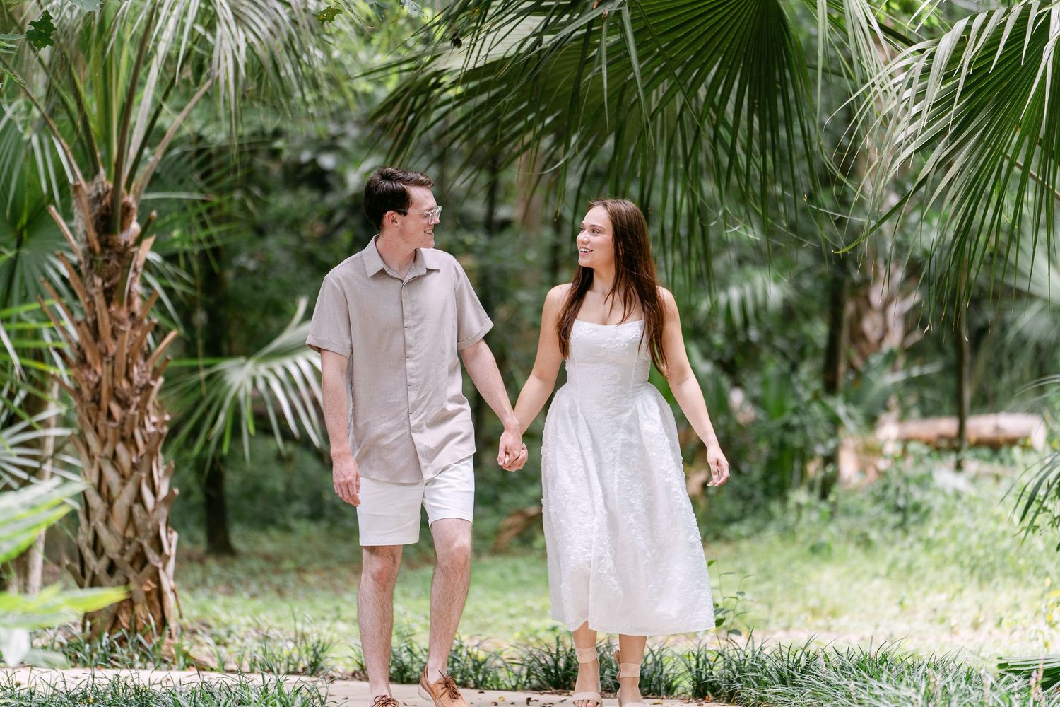 Couple holding hands walking through lush tropical garden, man in grey shirt and woman in white dress.