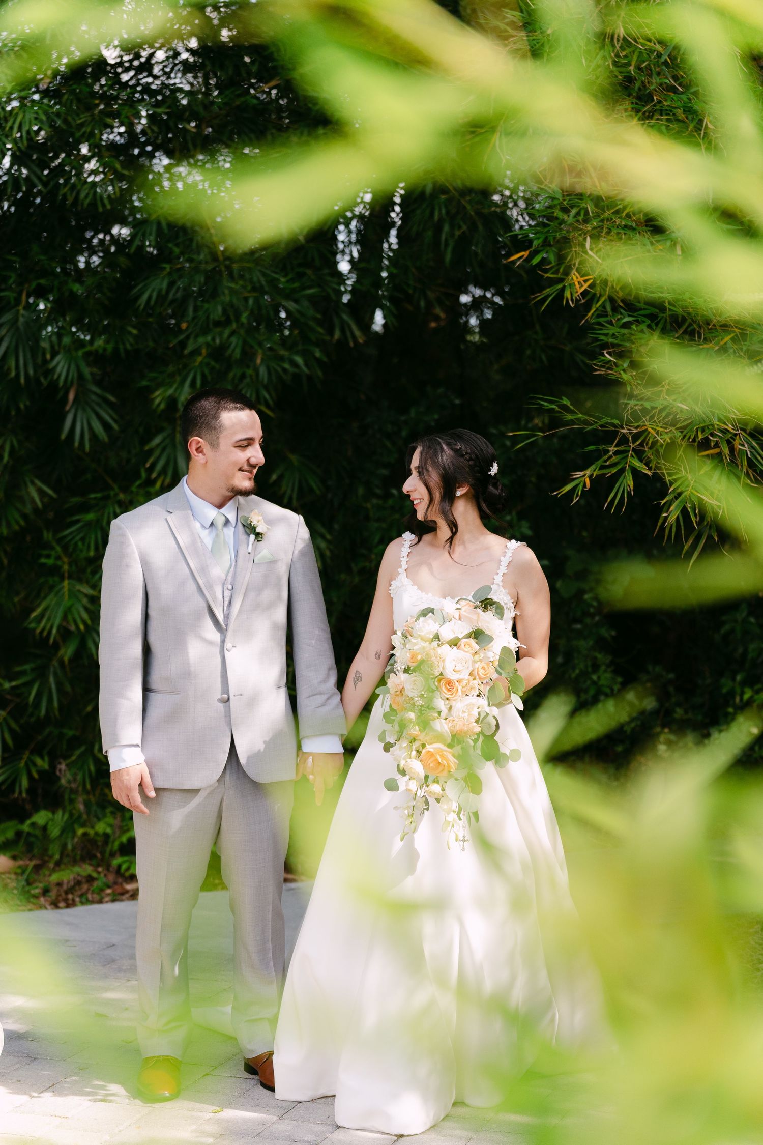 Bride and groom hold hands smiling in a lush garden, she holds a peach and white bouquet, he wears a light gray suit.