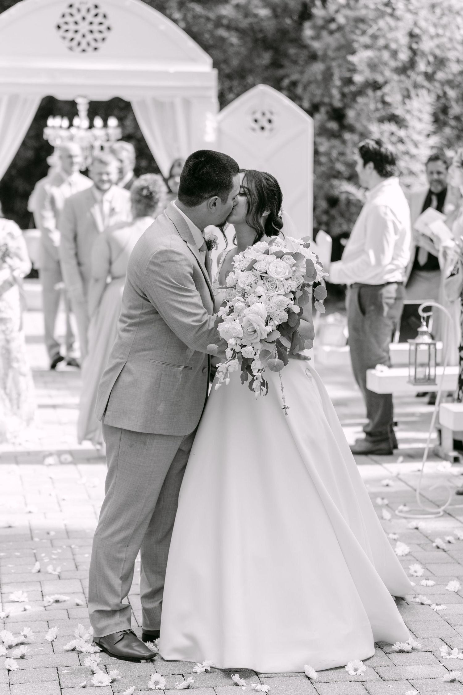 Black and white photo of bride and groom sharing first kiss at outdoor wedding ceremony with floral arch backdrop.