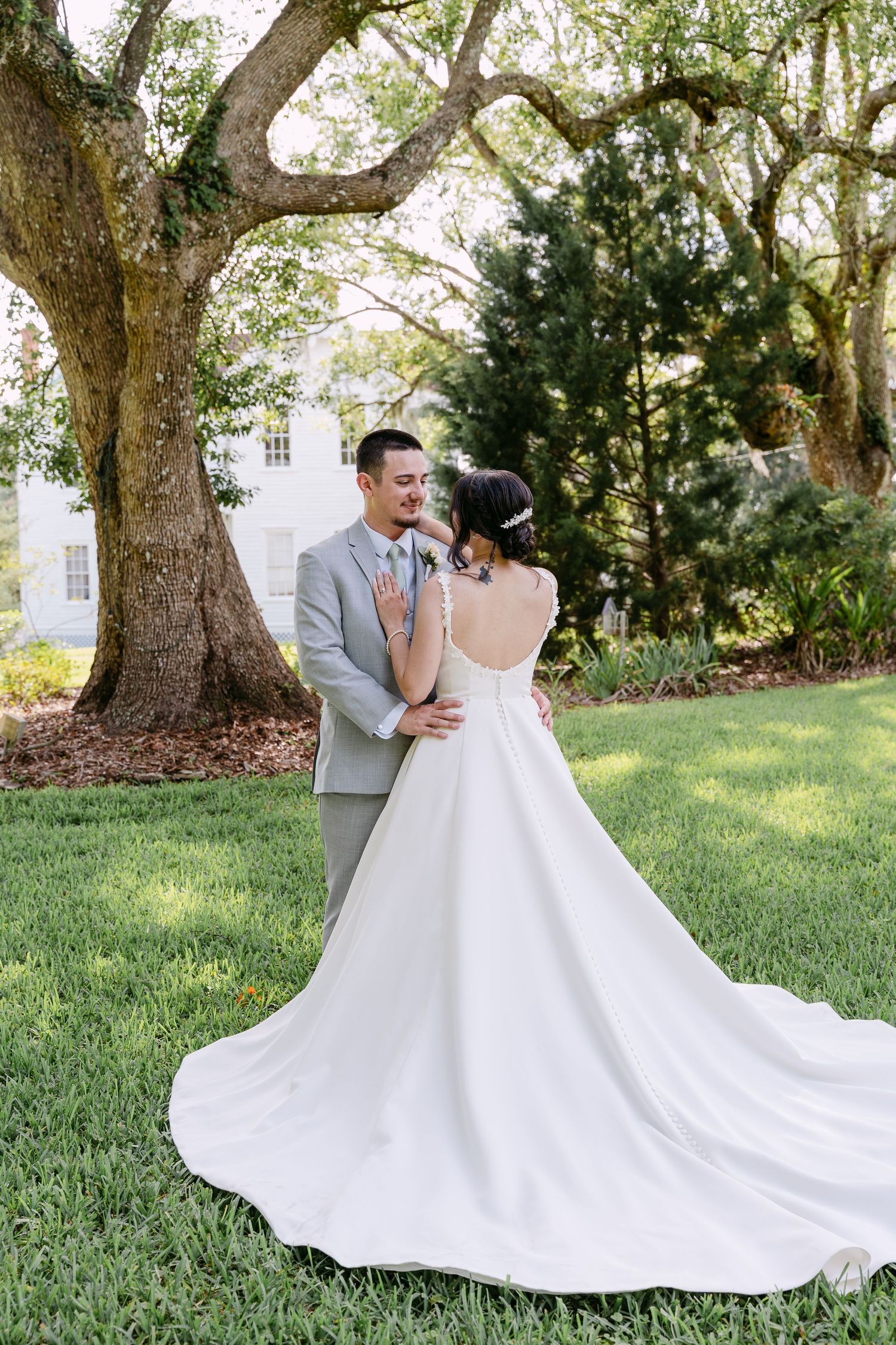 Bride and groom share a romantic moment on lush green lawn beside a grand oak tree at an elegant outdoor wedding.