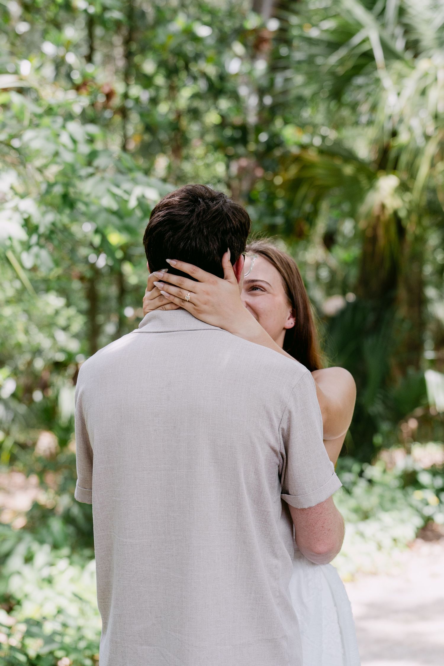 A couple shares a romantic embrace outdoors, surrounded by lush green tropical foliage.