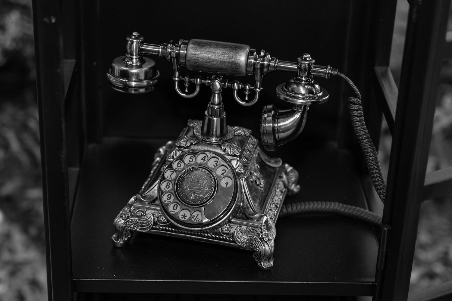 Black and white photo of an ornate vintage rotary telephone displayed on a dark shelf.
