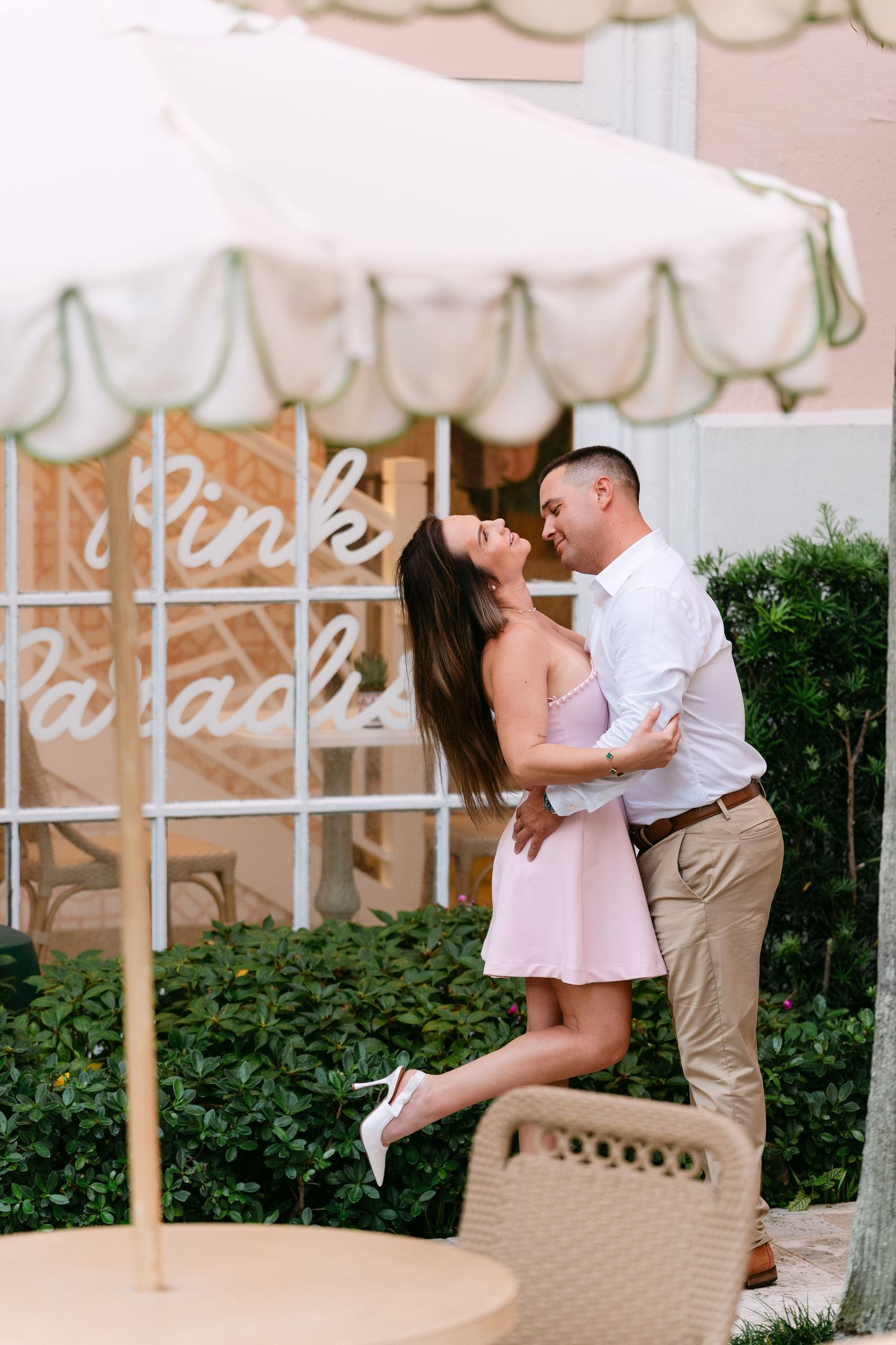 Couple sharing a romantic moment outside Pink Paradise, woman in pink dress lifted on tiptoes, under white striped umbrella.
