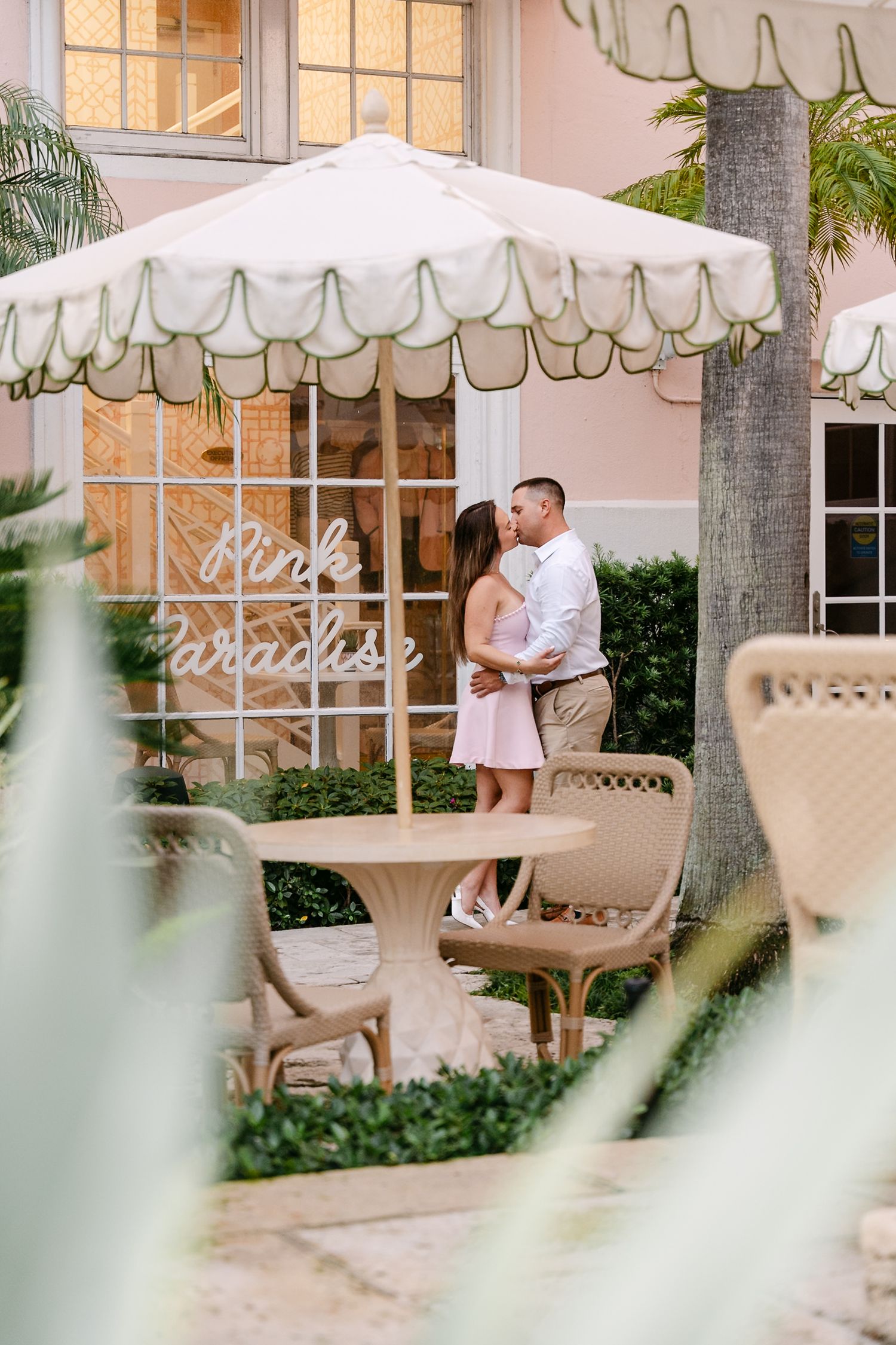 Couple kissing under a scalloped patio umbrella near a Pink Paradise neon sign in a garden setting.