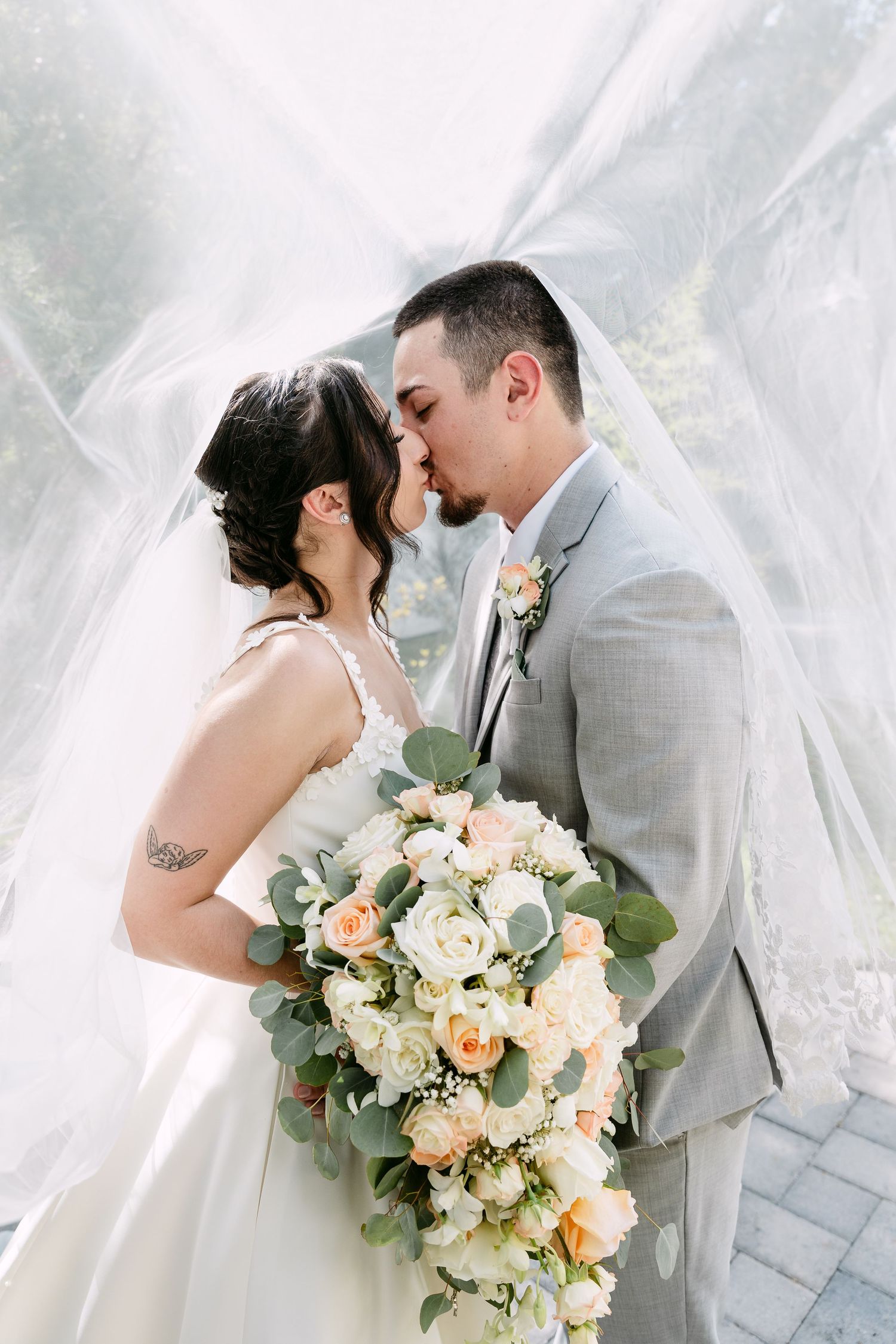 Bride and groom kissing under a flowing veil, bride holding a cascading bouquet of white and peach roses with eucalyptus.