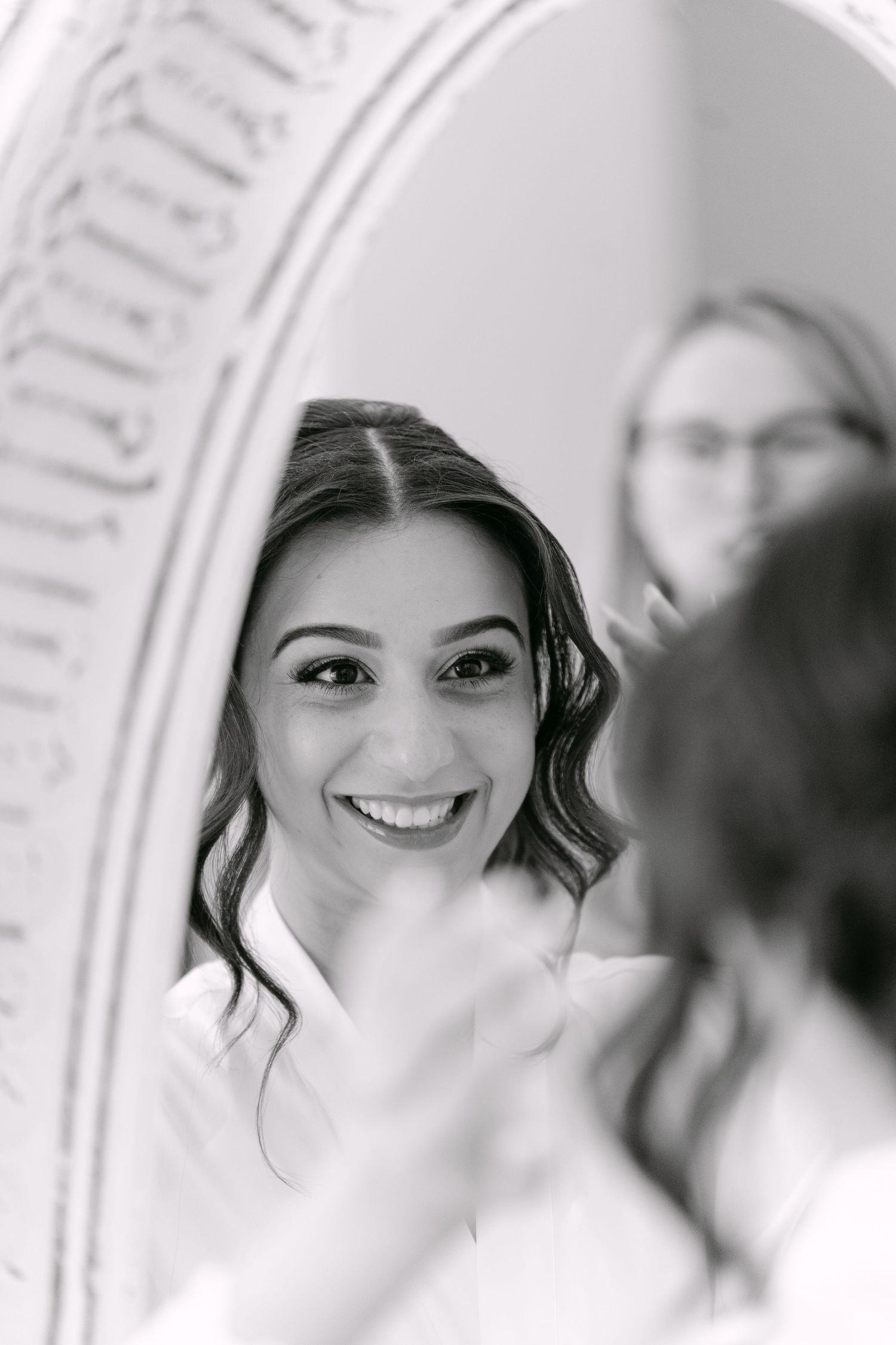 Black and white photo of a smiling bride getting makeup done, reflected in an ornate mirror.