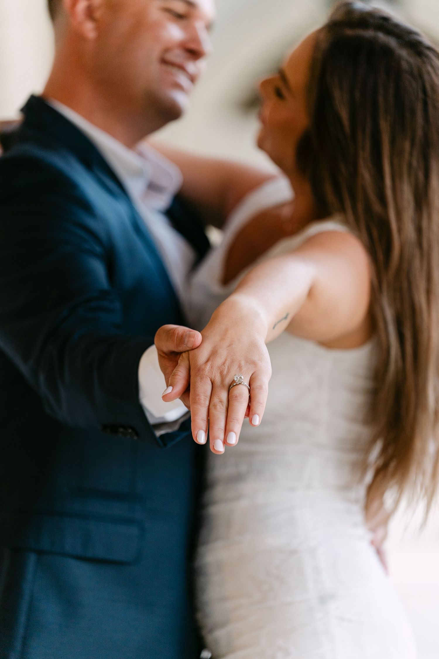 Smiling couple showing off a sparkling engagement ring while embracing in a bright indoor setting.