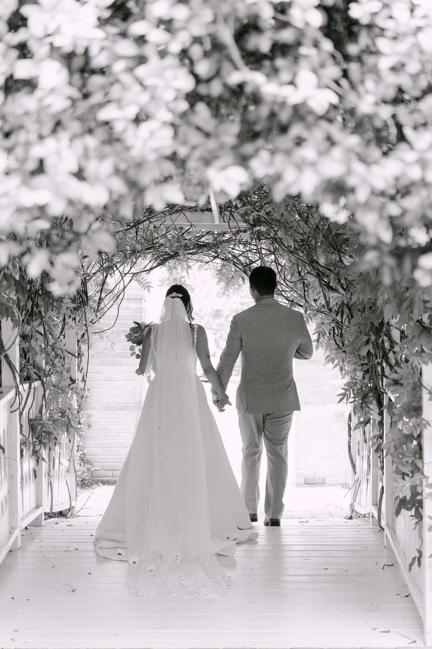 Black and white photo of bride and groom holding hands walking through a floral arch tunnel on their wedding day.