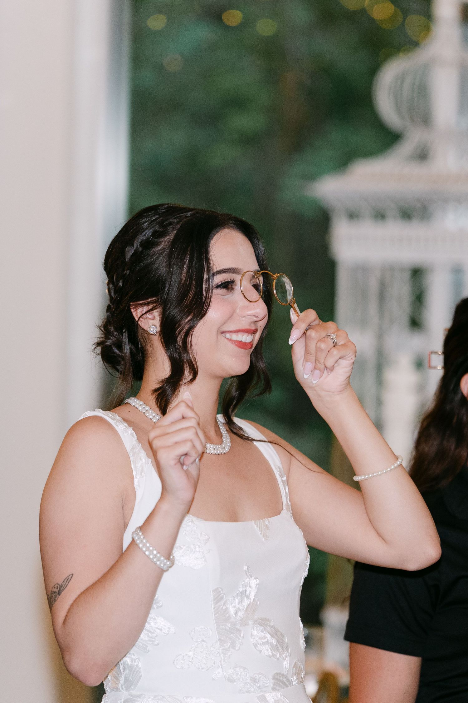 Smiling bride in white lace gown adjusting gold glasses, wearing pearl necklace and bracelet at elegant wedding venue.