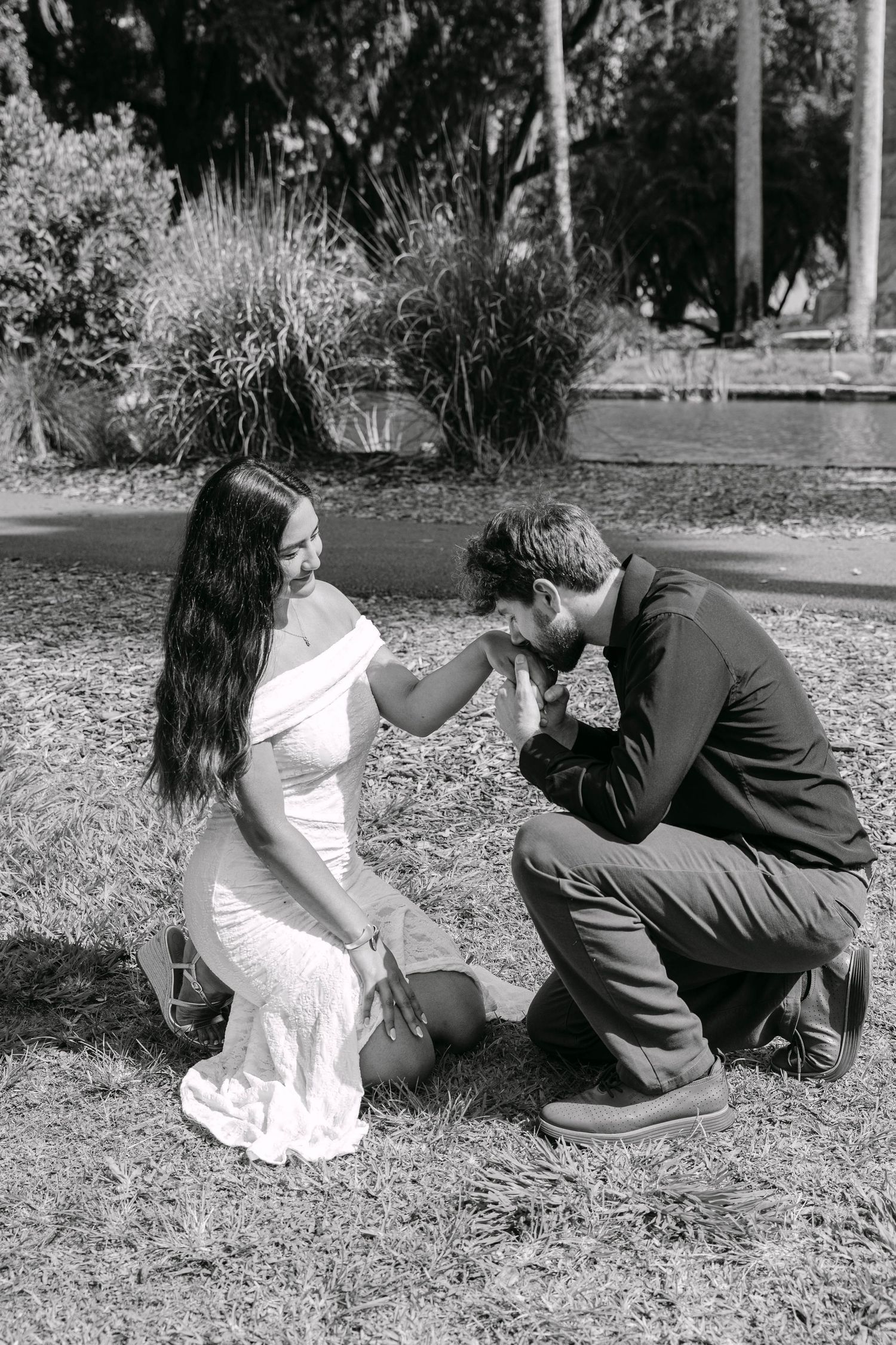 Black and white photo of man kneeling to kiss womans hand in park.