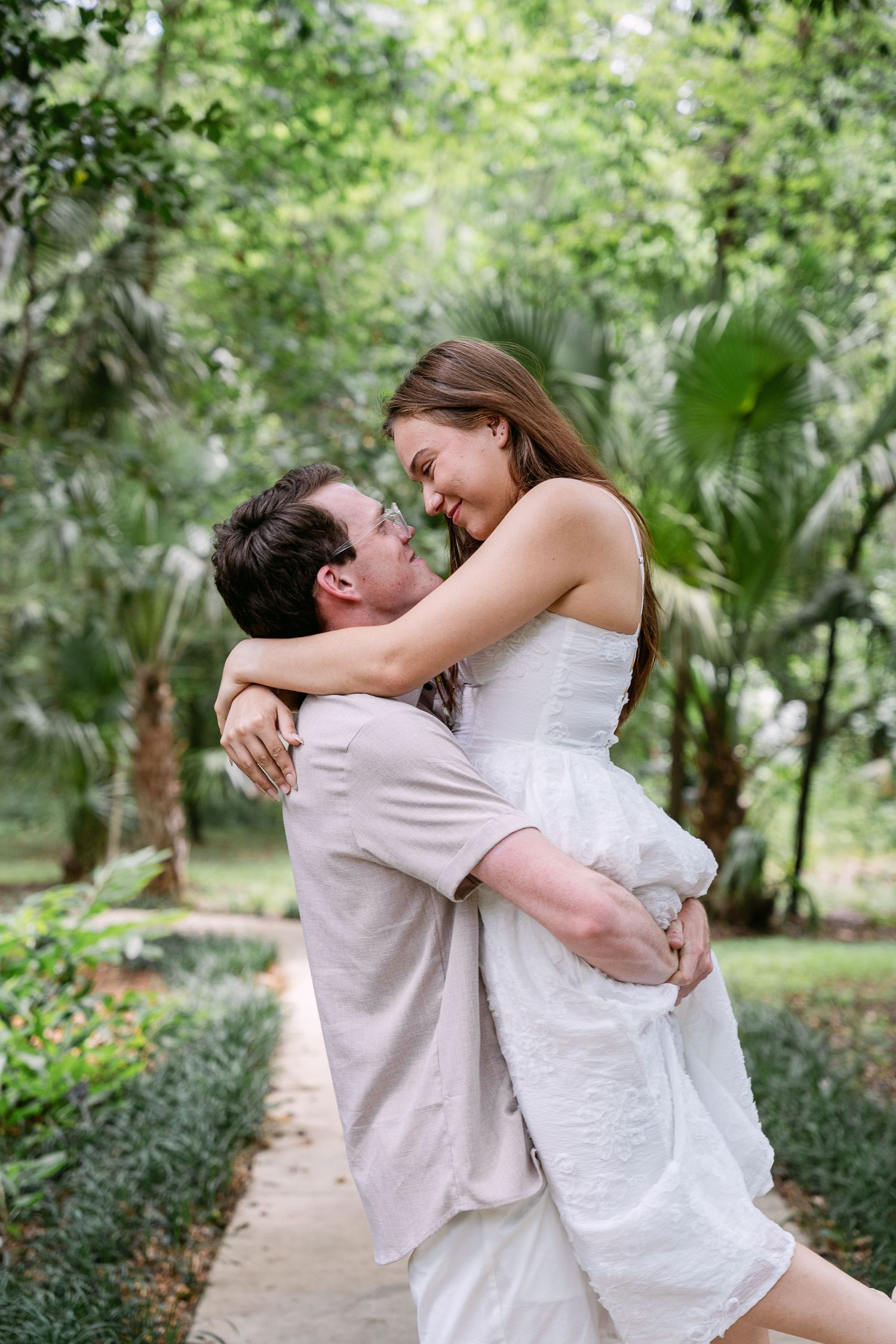 A joyful couple embraces in a lush green garden, the man lifting the woman in a white dress as they smile at each other.