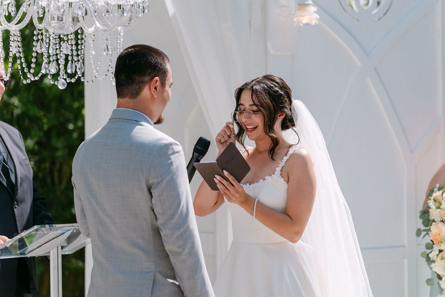 Bride in white wedding gown reads vows while smiling at groom in gray suit during outdoor wedding ceremony.
