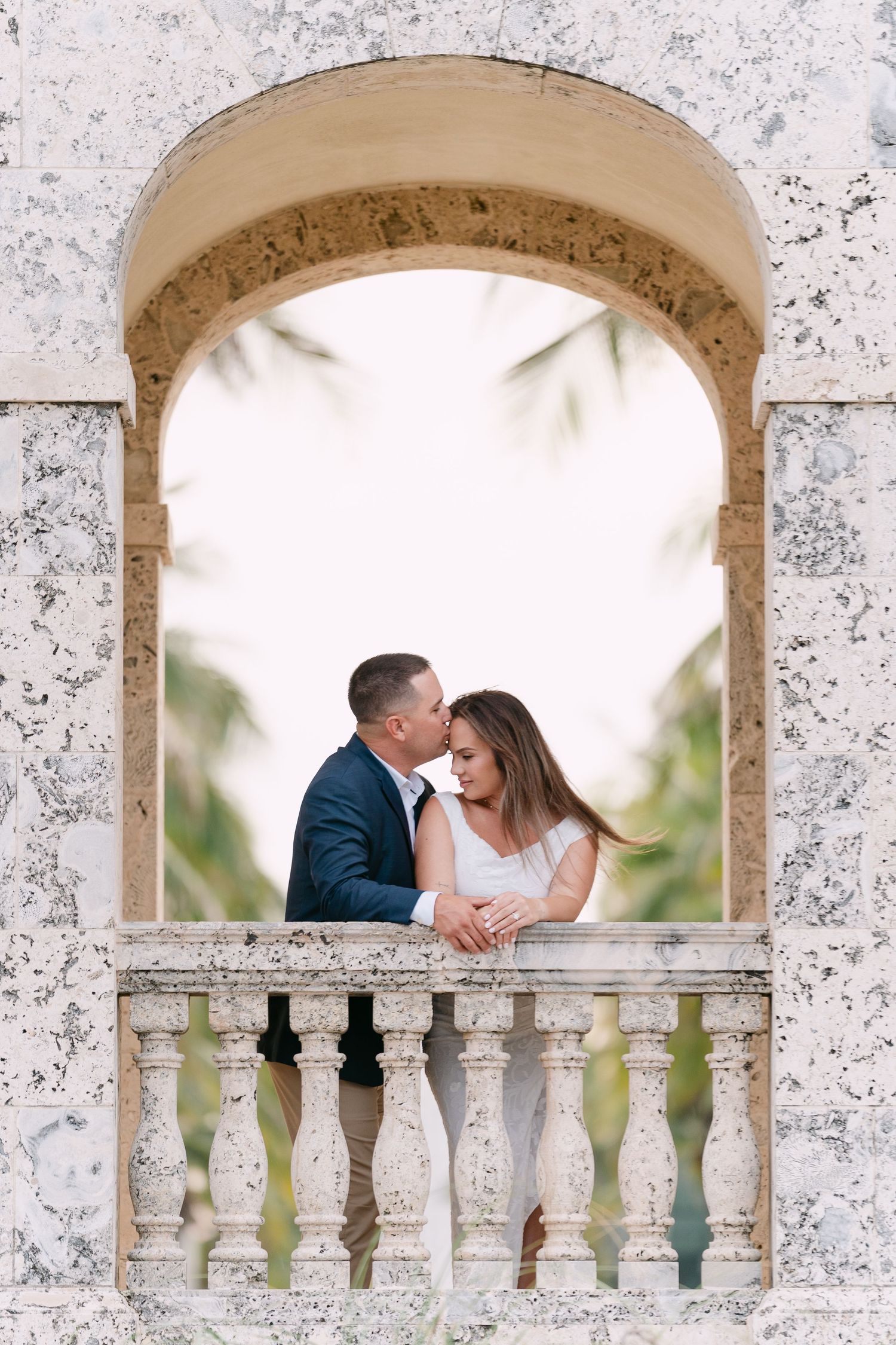 Couple sharing a romantic kiss on an ornate marble balcony framed by an elegant stone arch with palm trees.
