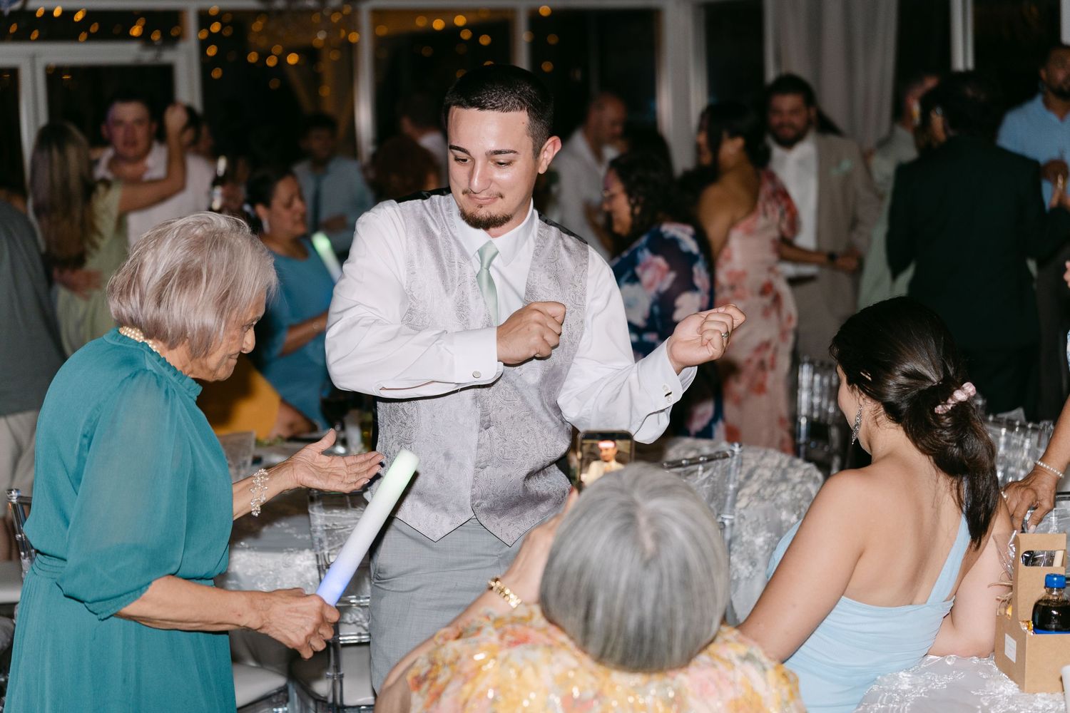 Groom in gray suit dancing and greeting elderly guests at a wedding reception with guests seated at tables.