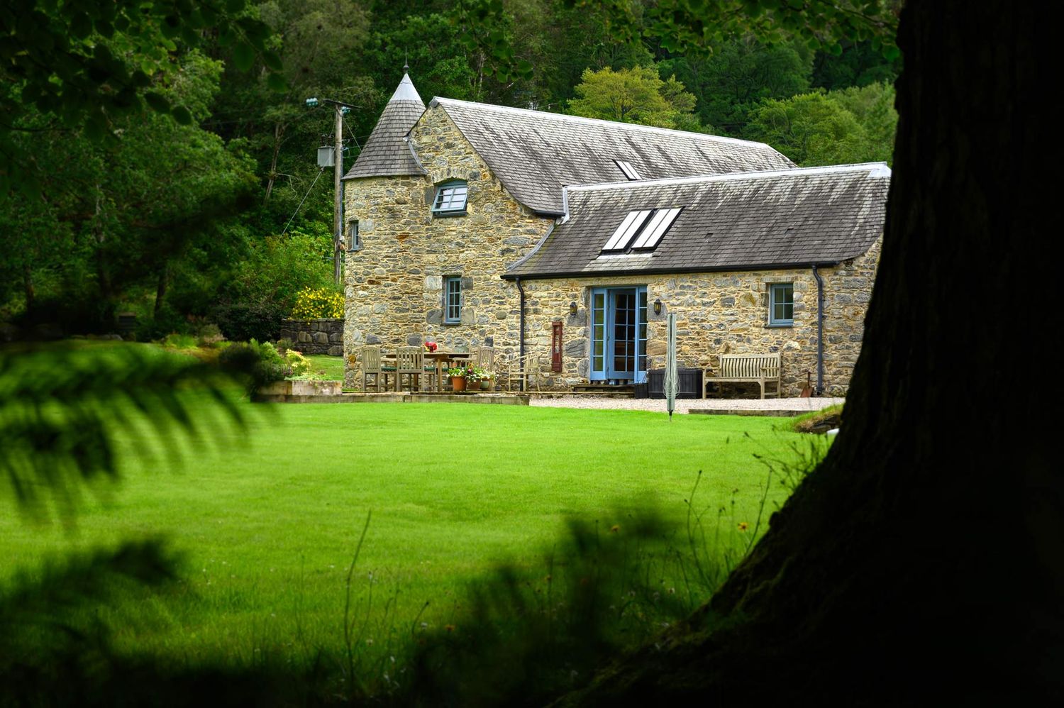 Exterior view of a stone cottage with a large green lawn framed by trees.