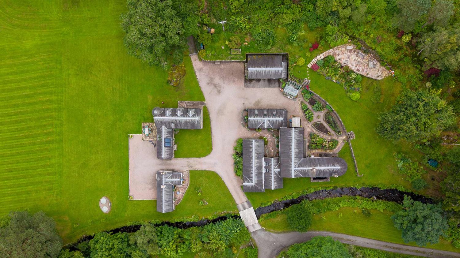 Top-down aerial view of a stone cottage complex with surrounding grounds, gravel paths, and a stream.
