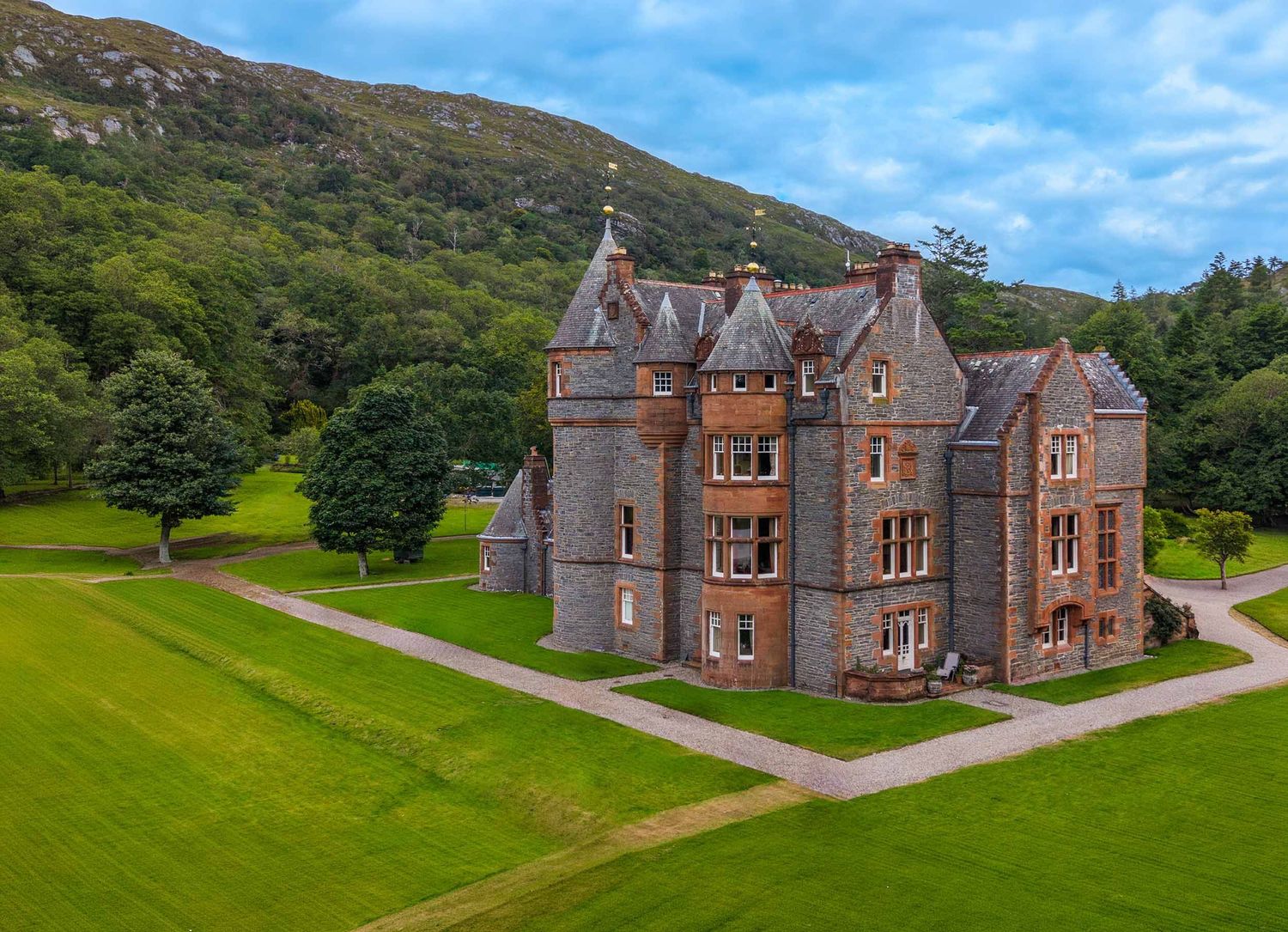 Elevated drone view of a turreted stone country house with lawns and a wooded hillside in the Scottish Highlands.