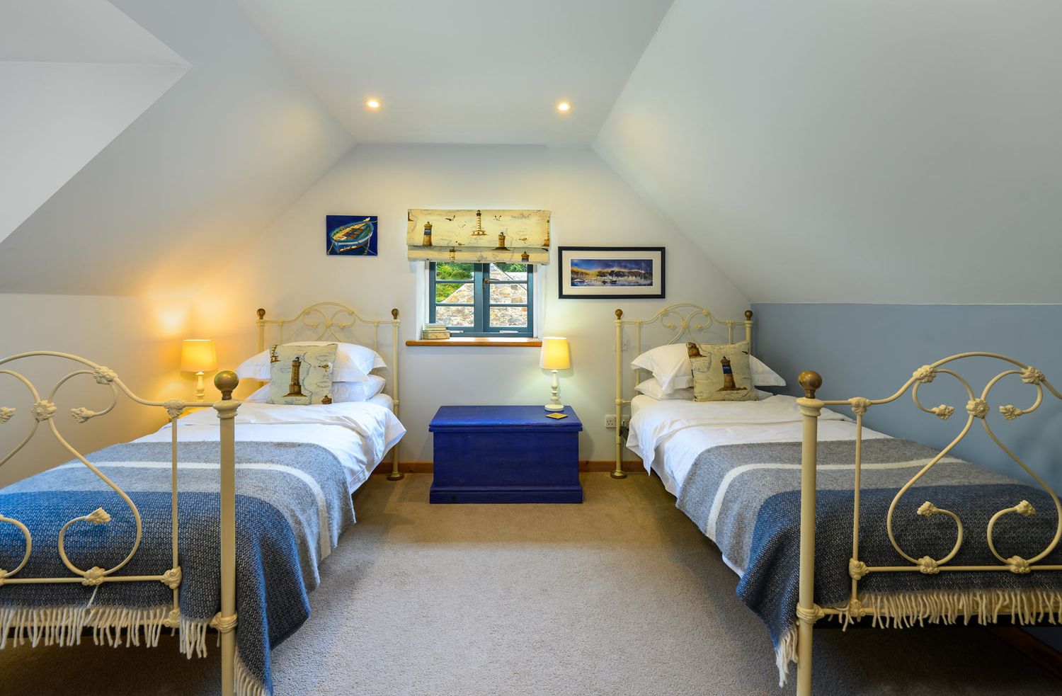 Cosy nautical themed bedroom featuring two metal frame twin beds separated by a blue chest and central window with ambient lighting.