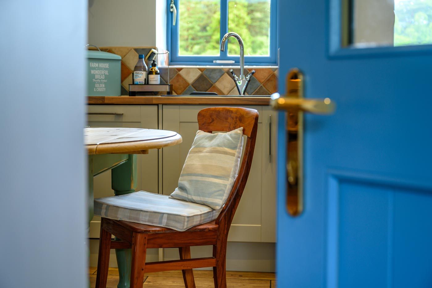 View through a bright blue open doorway into a cottage kitchen, showing a dining chair with a striped cushion and a sink.