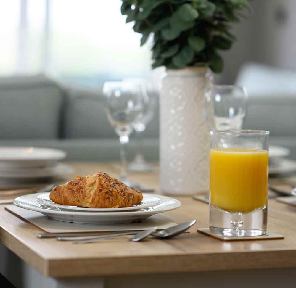 A styled breakfast table featuring a croissant on a white plate, a glass of orange juice, wine glasses, and a vase with greenery.