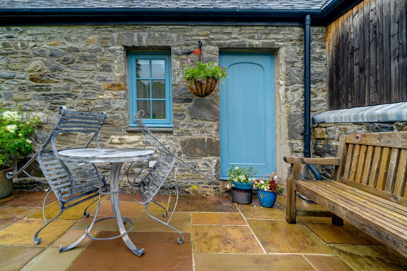 A rustic stone cottage courtyard with a flagstone patio, featuring a metal bistro table and chairs, a wooden bench, and a matching blue door