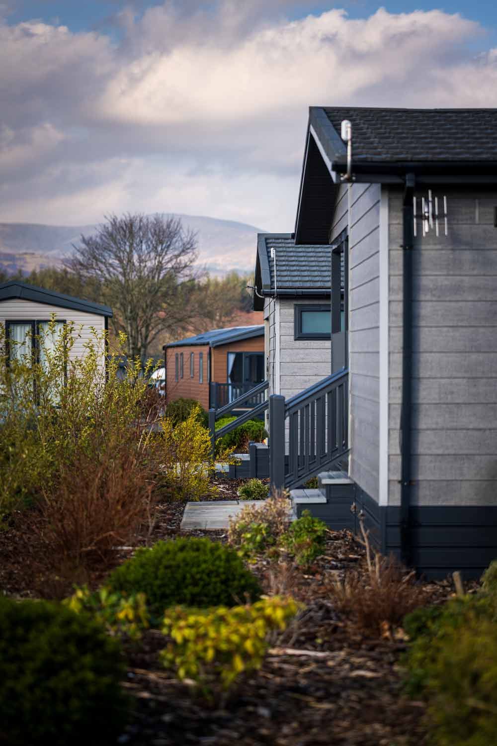 Modern grey holiday lodges situated in a landscaped setting with green shrubs, under a cloudy blue sky with distant hills.
