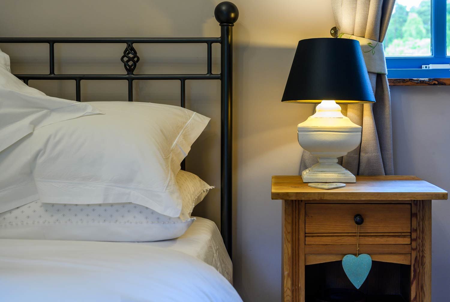 Cosy bedroom detail of a wooden bedside table with a glowing white lamp, black lampshade, and a decorative blue heart.