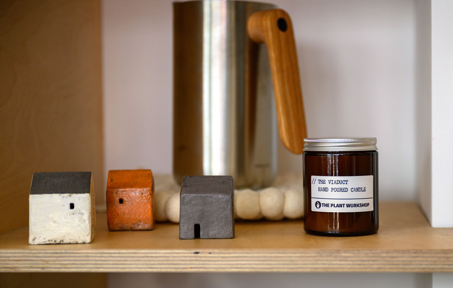 Close-up of wooden shelf decor featuring miniature house blocks, a metallic french press, and a scented jar candle.