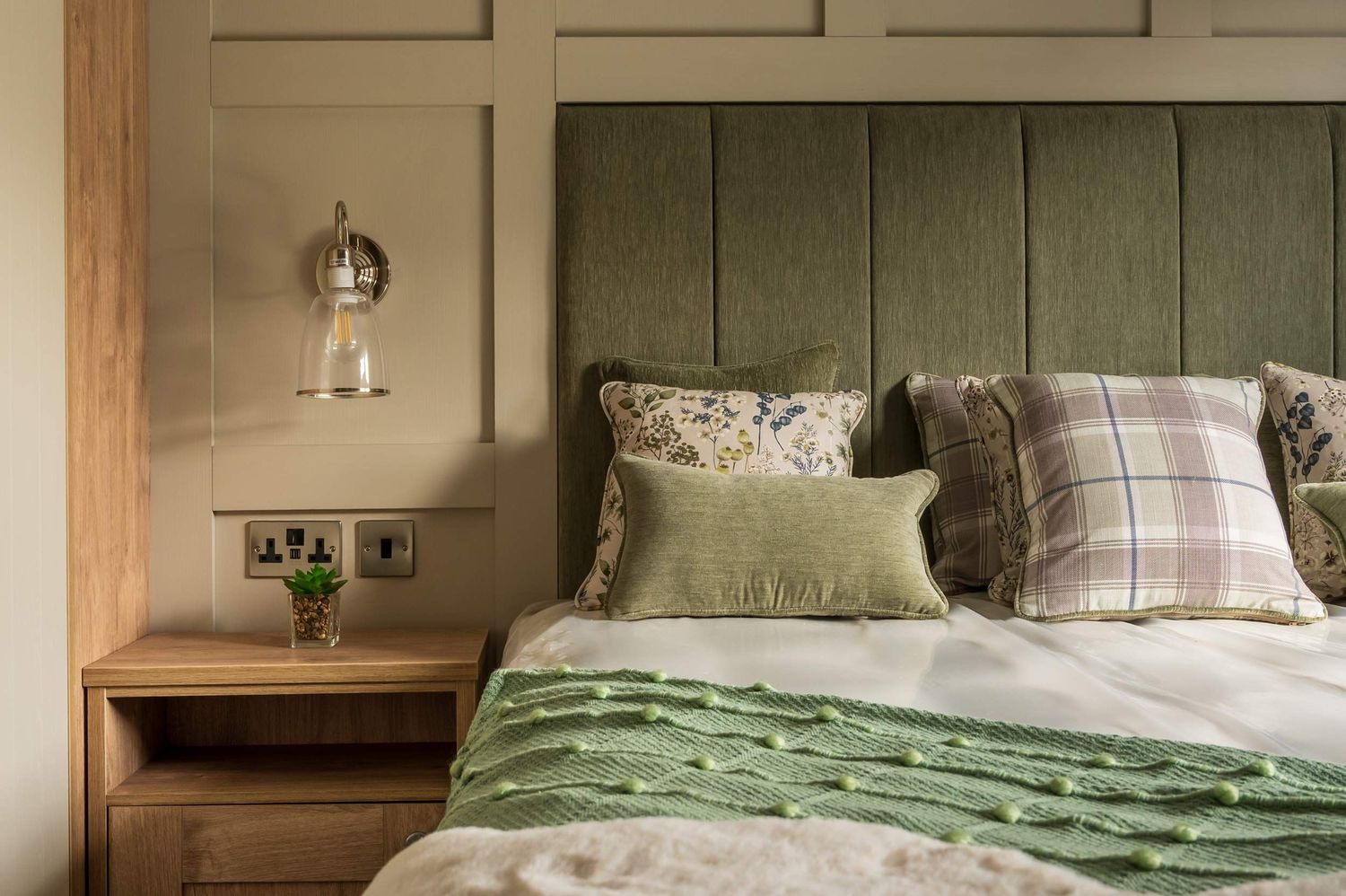 A styled bedroom featuring a sage green fluted headboard, plaid and floral cushions, a green throw, and a wooden bedside table.