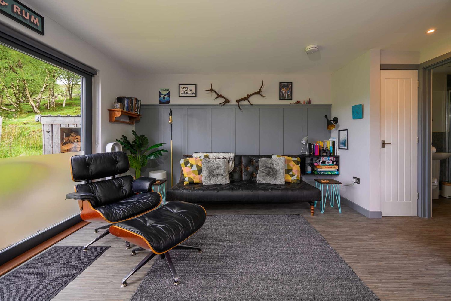 Modern living room with black leather lounge chair and sofa, grey panelled feature wall, bookshelves, decorative antlers, and a large window
