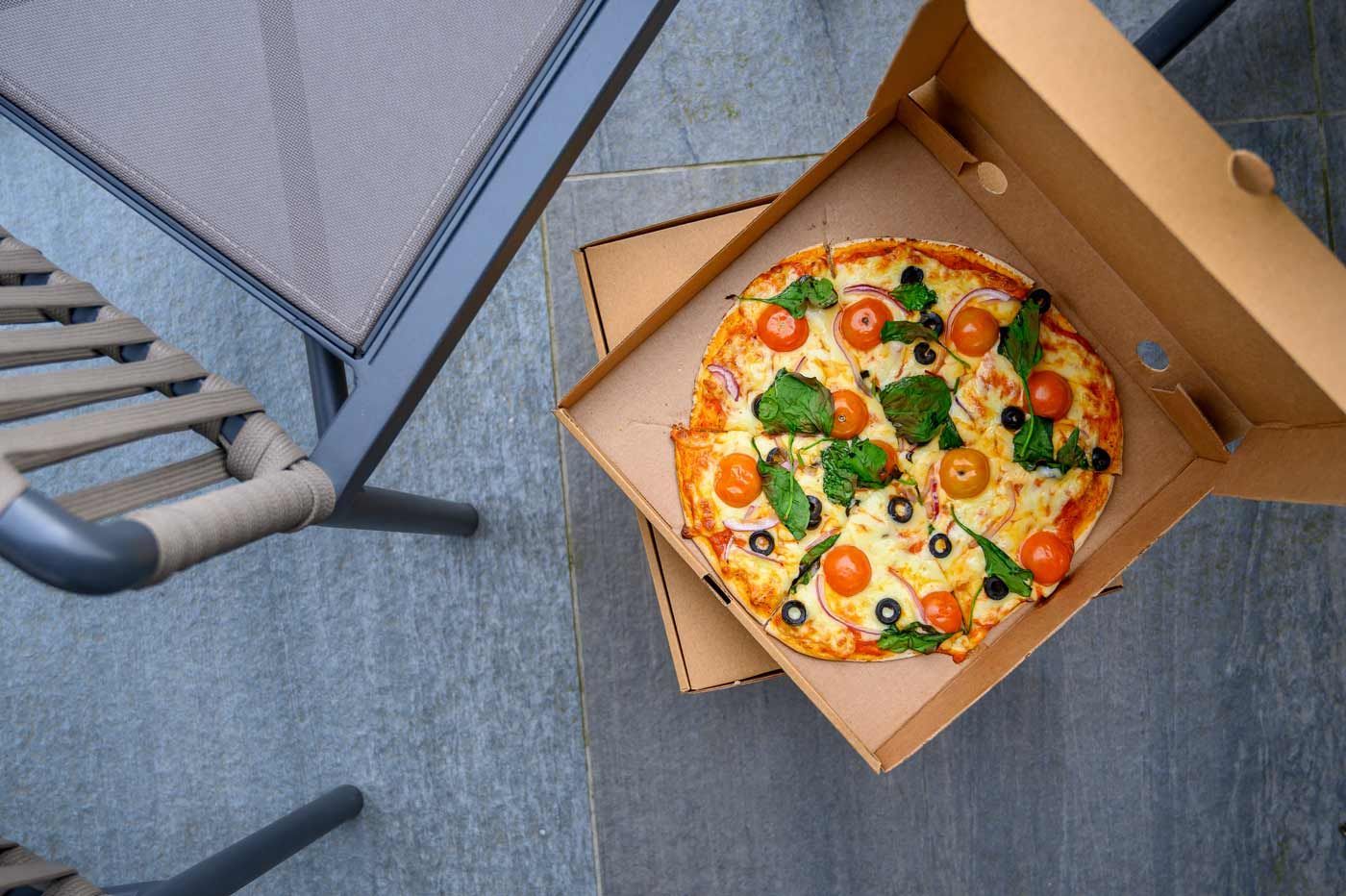 An overhead view of a fresh tomato and basil pizza in a cardboard delivery box, sitting on a patio floor next to a metal chair.