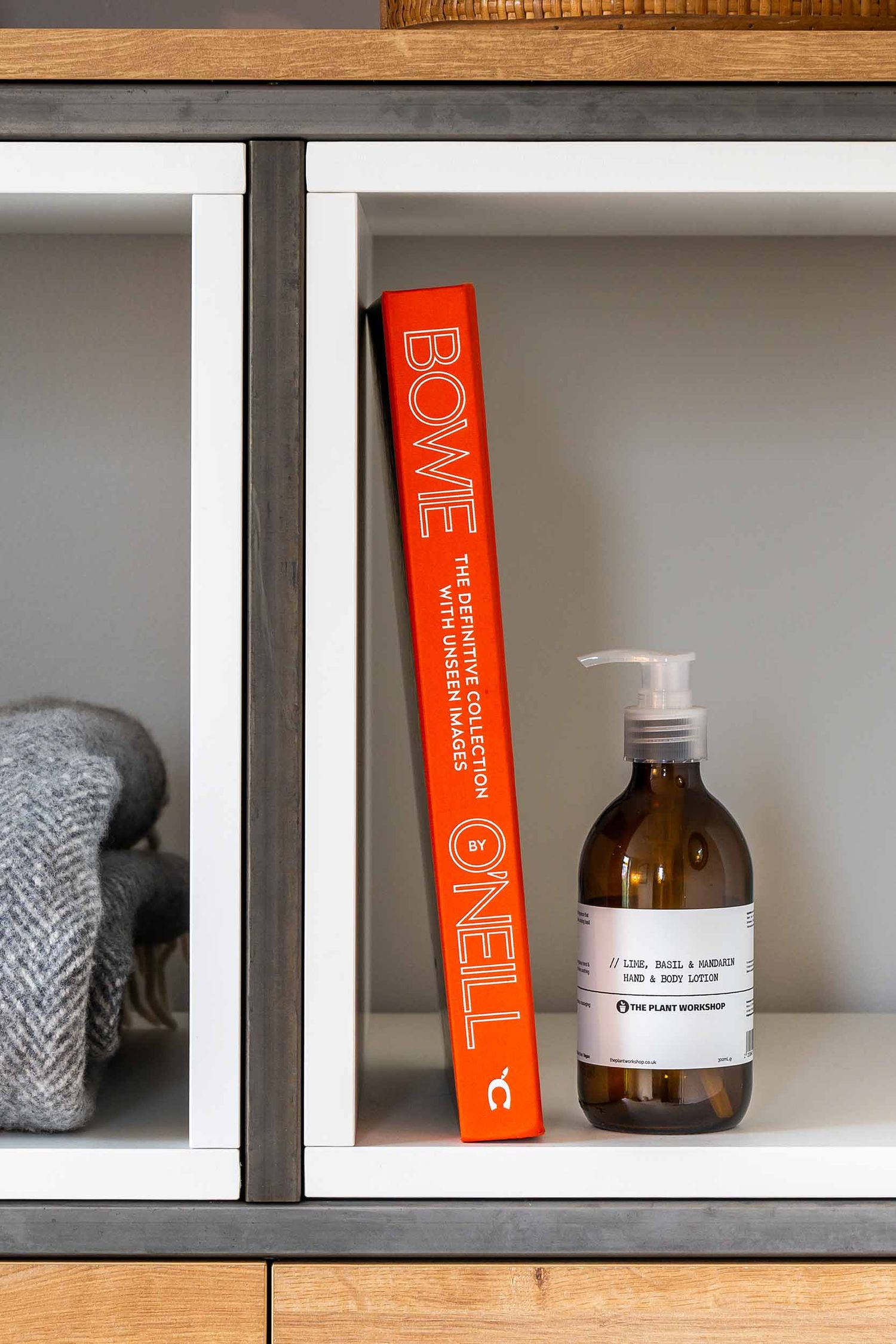 An amber glass hand lotion bottle sitting next to a bright orange David Bowie book on a white shelving unit.