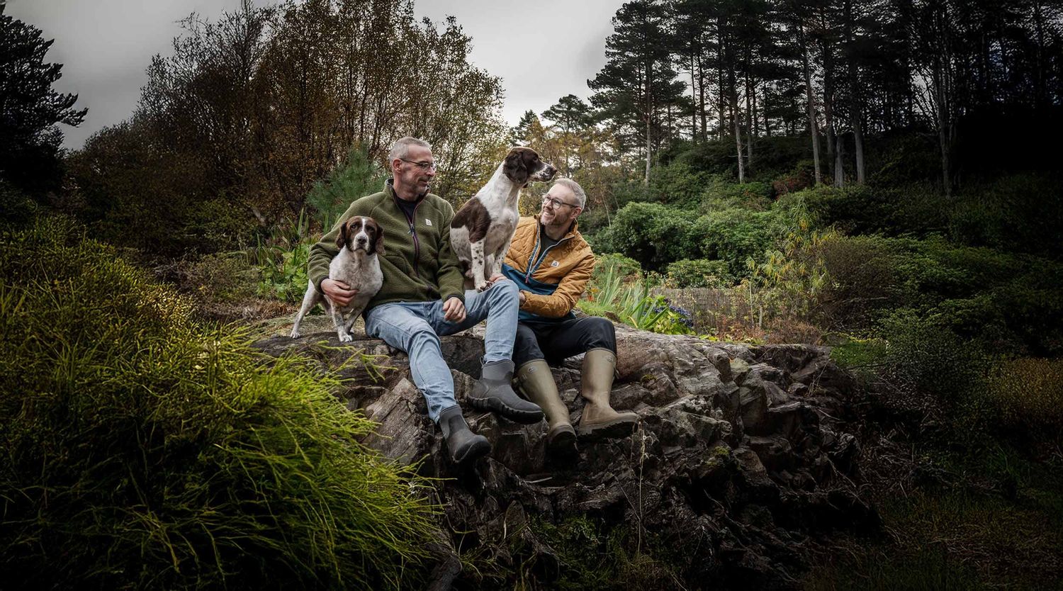 An outdoor portrait of Dave & family sitting on a mossy stone wall in a wooded area with two spaniel dogs.
