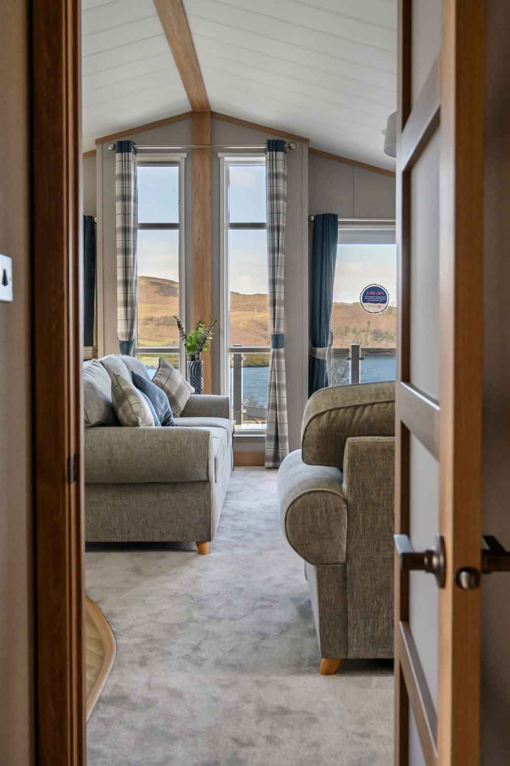 A view through an open wooden doorway looking into a bright, modern living room with large windows showing a coastal landscape.