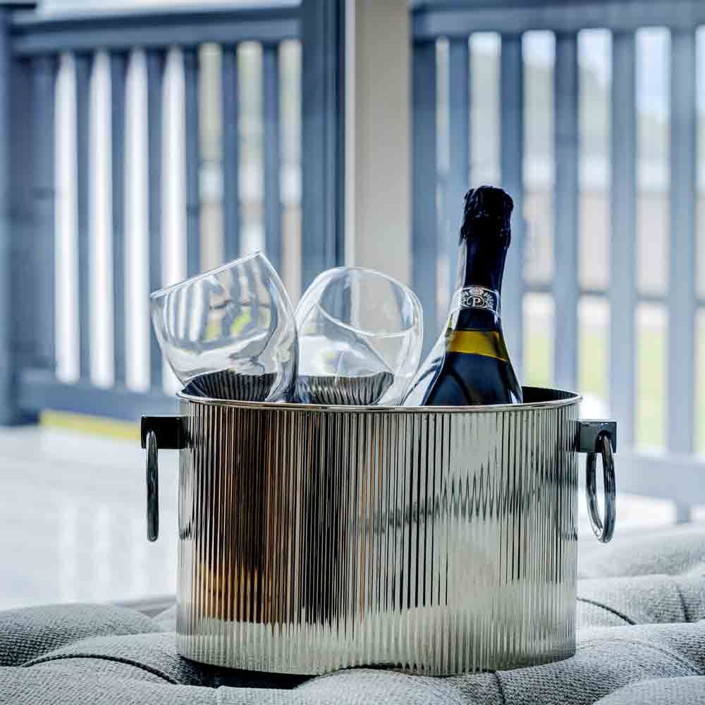 A silver champagne bucket containing a bottle of wine and two glasses, resting on an outdoor tufted footstool with a deck railing.