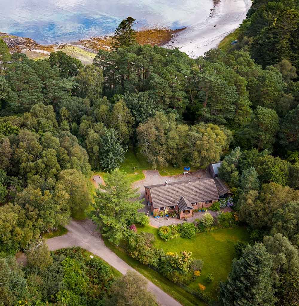 Aerial drone view of a cottage surrounded by woodland and gardens beside a sandy shoreline in the Scottish Highlands.