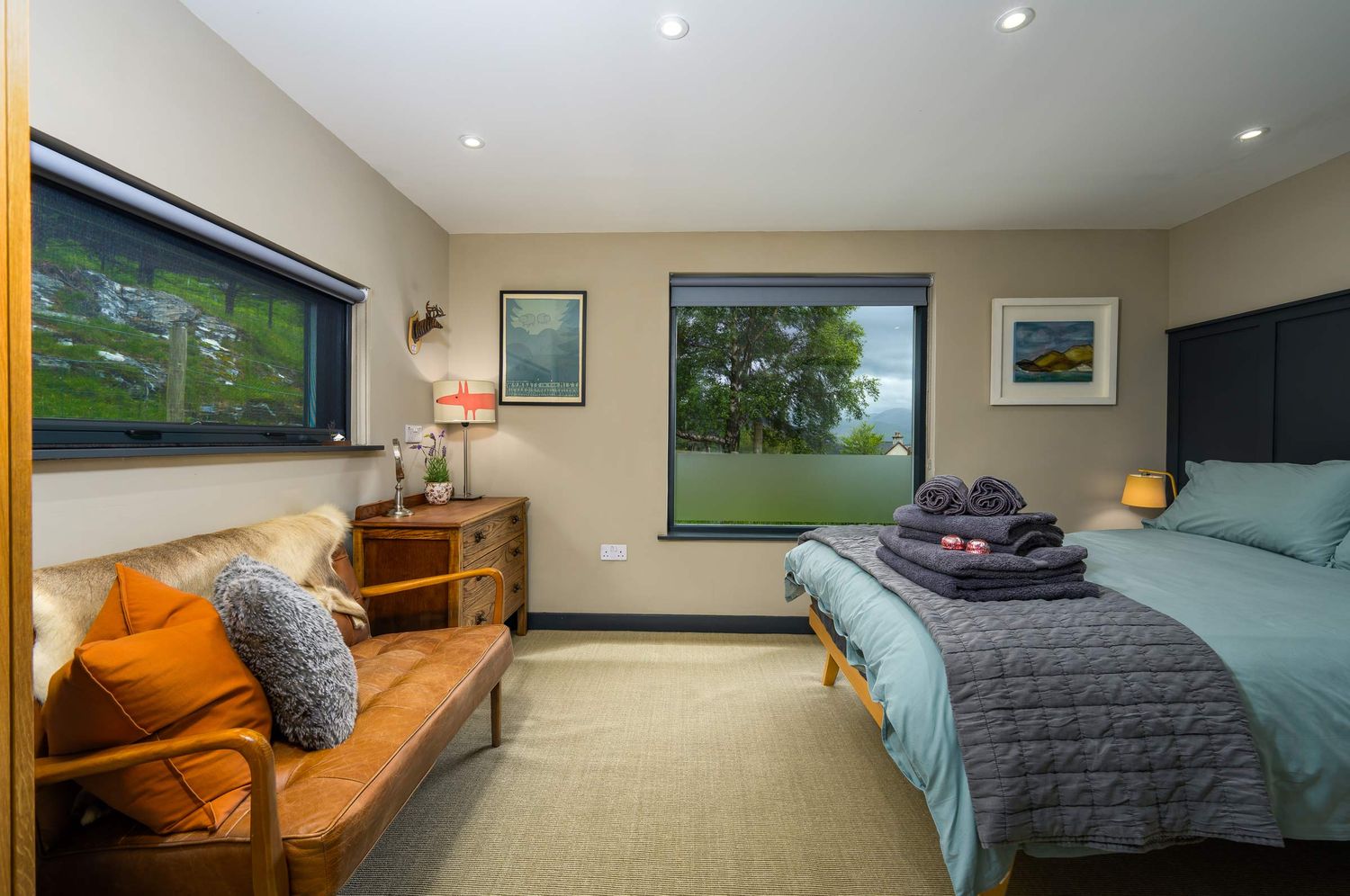 Spacious cottage bedroom featuring a double bed with blue linens, a tan wooden bench with orange cushions, and bright windows.