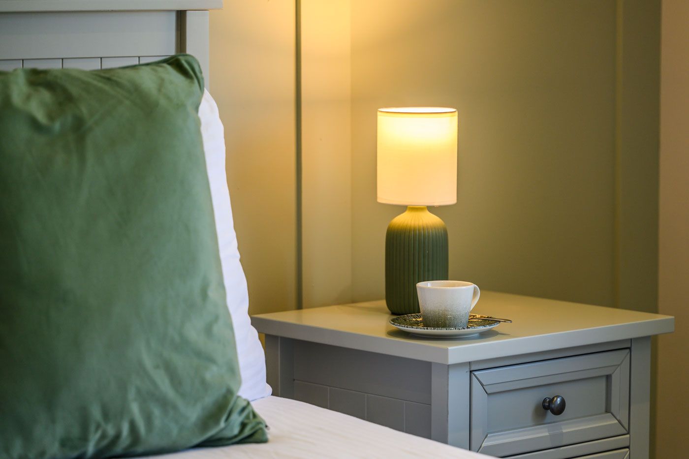 A bedside view featuring a glowing table lamp, a white coffee cup on a saucer, and a large green pillow on the bed.