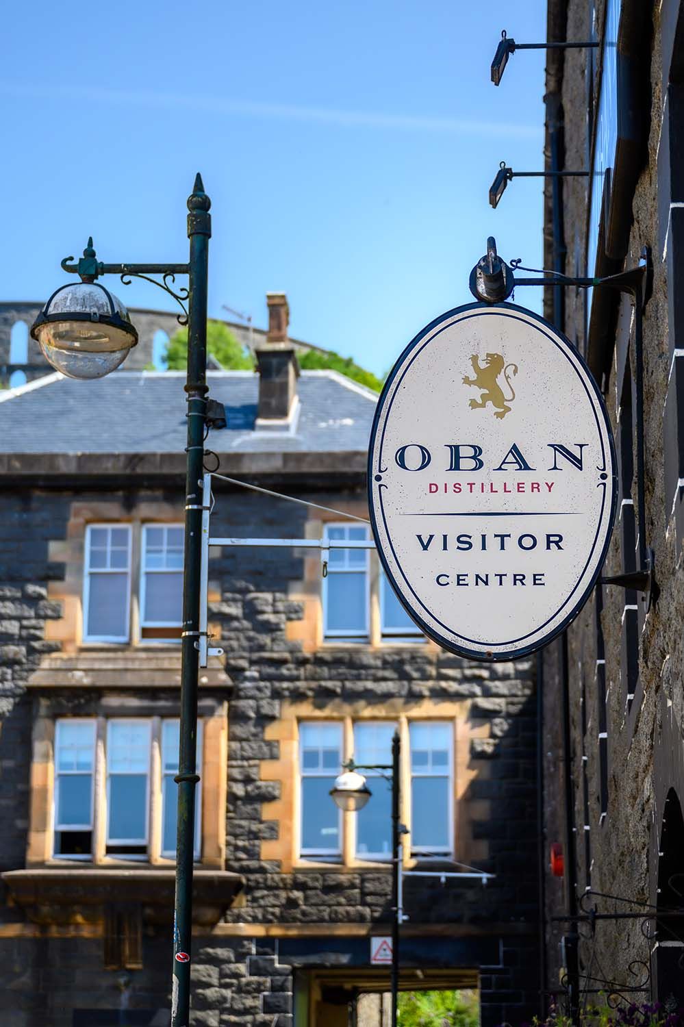 Exterior sign for Oban Distillery set against traditional stone buildings under a blue sky.