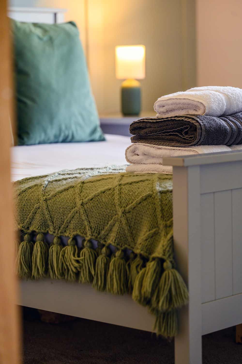 A wooden bed frame styled with a green tasselled blanket, a stack of towels, a green pillow, and a glowing bedside lamp.