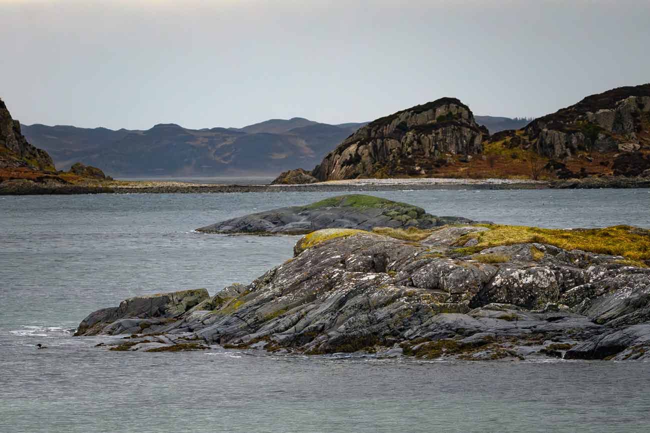 Rocky, moss-covered outcrops along a coastline with calm sea waters and rolling hills in the background.