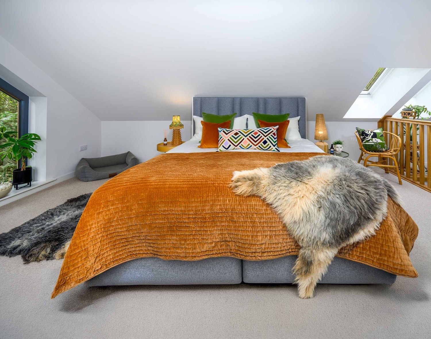 Modern loft bedroom featuring a grey fabric bed dressed with a rust-orange velvet quilt, colourful cushions, and a sheepskin throw.
