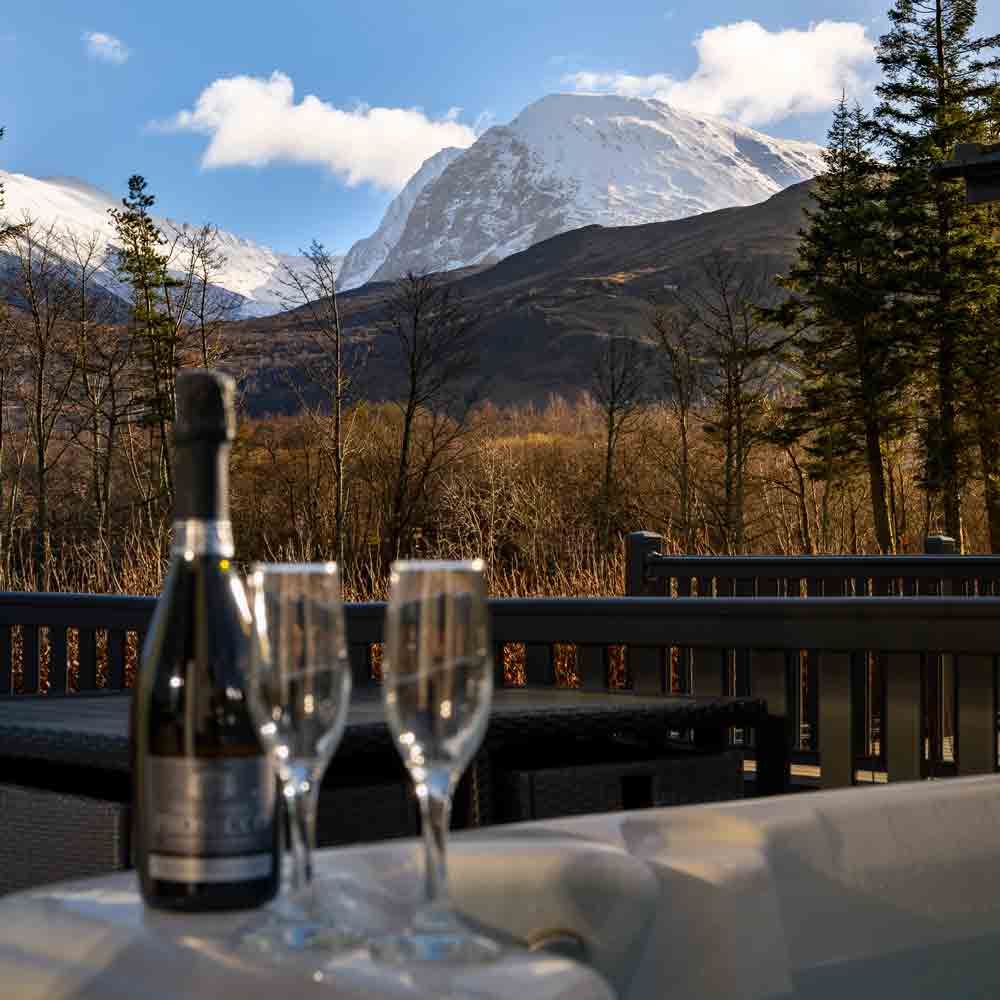 A bottle of bubbly and two glasses sitting by a hot tub, overlooking a wooden deck with a dramatic, snow-capped mountain behind.