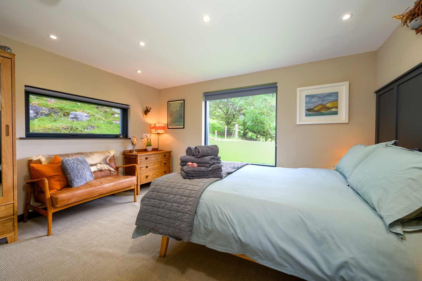 Spacious bedroom with a light blue bed, a tan leather bench under a panoramic window, and bright countryside views.