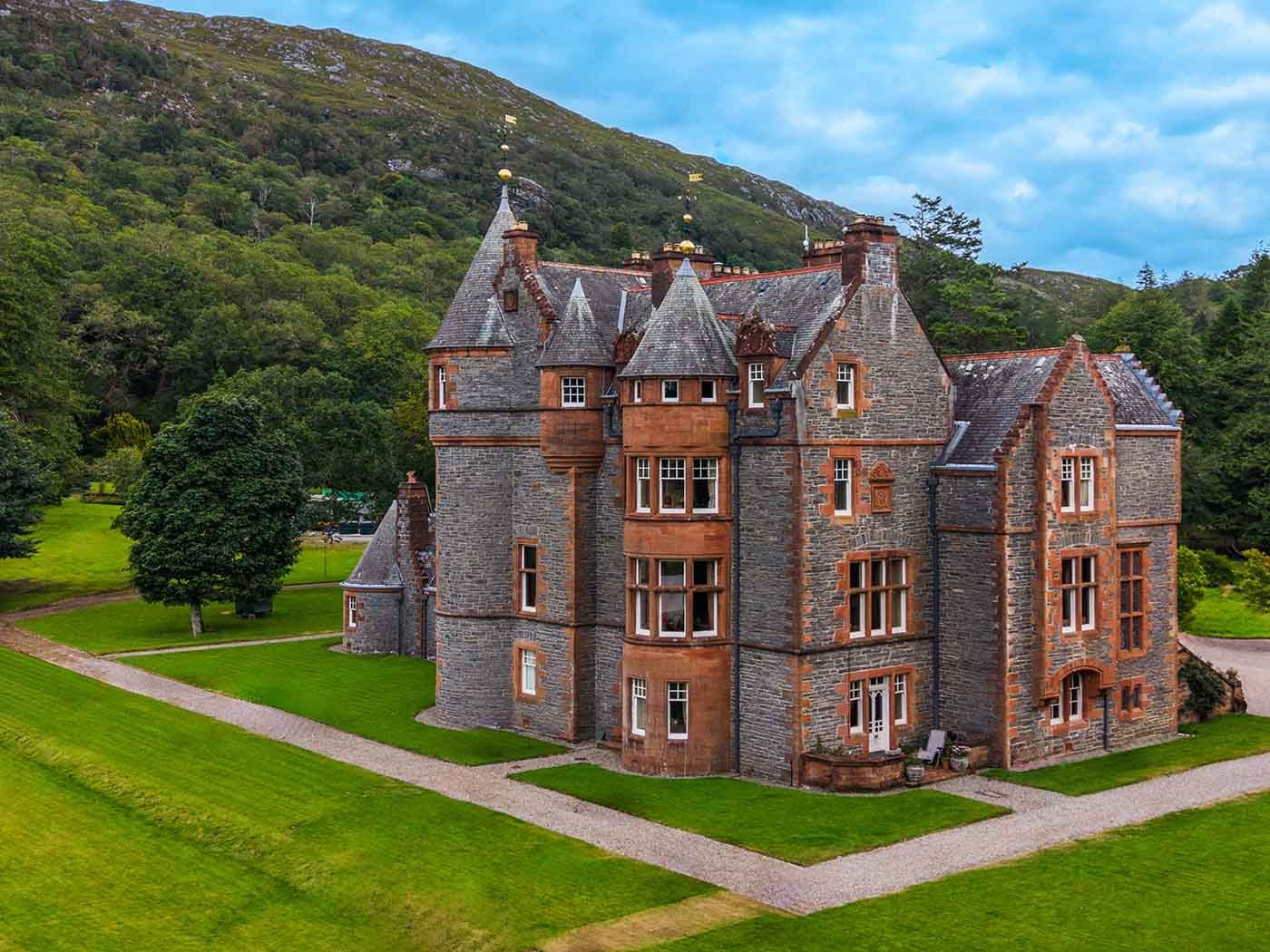 Elevated drone view of a turreted stone country house surrounded by lawns and woodland in the Scottish Highlands.