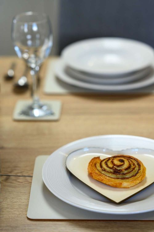 A table setting featuring a cinnamon swirl pastry on a heart-shaped plate, a wine glass, and stacked dinnerware.