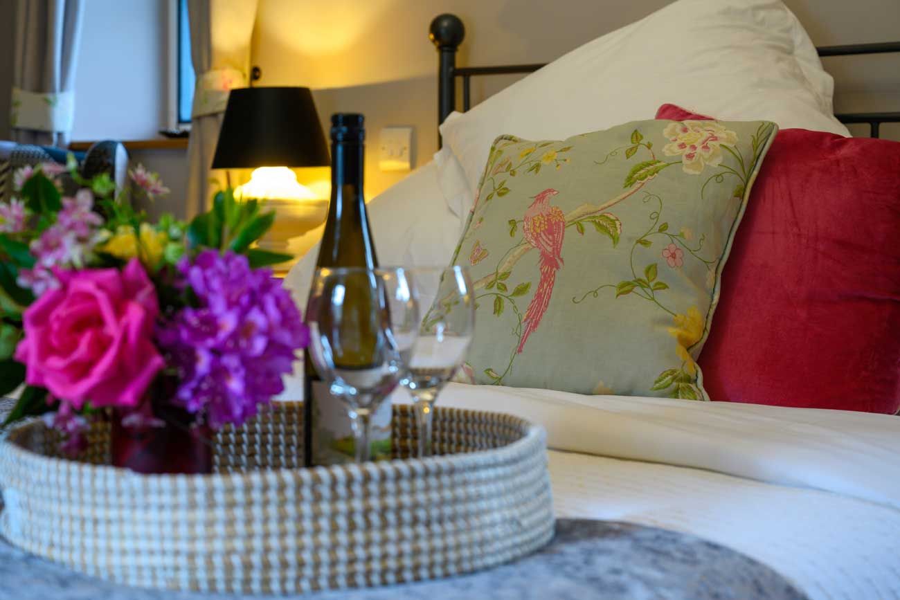 A welcome basket on a bed with fresh pink peonies in a vase, a bottle of wine, two glasses, and a floral accent pillow.